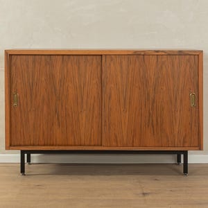 May include: A mid-century modern wooden cabinet with two sliding doors. The cabinet is made of a warm-toned wood with a visible grain pattern. It has brass handles and sits on black metal legs.