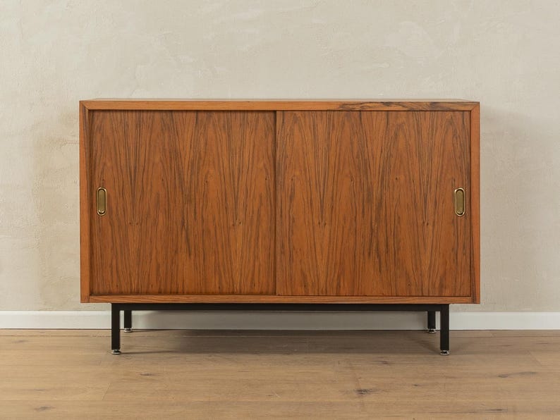 May include: A mid-century modern wooden cabinet with two sliding doors. The cabinet is made of a warm-toned wood with a visible grain pattern. It has brass handles and sits on black metal legs.