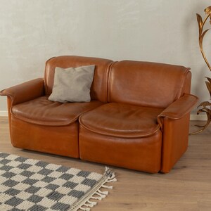 Könnte beinhalten: Ein Zweisitzer-Ledersofa in Braun mit Armlehnen. Ein graues Kissen liegt auf dem linken Sitz. Das Sofa steht auf einem Holzboden, im Vordergrund liegt ein karierter Teppich. Im Hintergrund befindet sich eine goldfarbene Stehlampe. Das Sofa ist ca. 180 cm breit.
