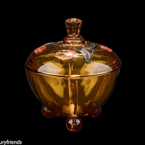 May include: Amber glass candy dish with a lid, decorated with black and red floral designs. The dish has three feet.