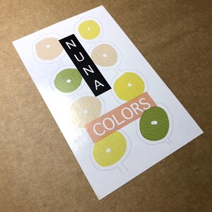 Peut inclure: Une carte blanche avec un motif color&eacute;. La carte pr&eacute;sente une s&eacute;rie de cercles dans diff&eacute;rentes nuances de jaune, de vert et de beige. Le mot "NUNA" est imprim&eacute; en noir sur la carte, et le mot "COLORS" est imprim&eacute; en rose.