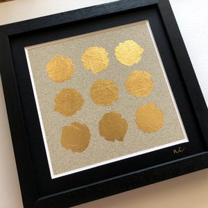 May include: A framed artwork with a beige background and 10 gold circles arranged in a grid pattern. The artwork is signed "nc" in the bottom right corner.