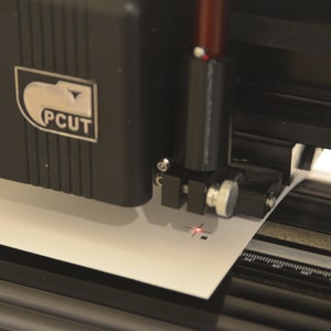 May include: A close-up of a black PCUT vinyl cutter with a red laser pointer, cutting a white piece of paper.