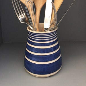 May include: A blue and white striped ceramic utensil holder with a wooden spatula, two wooden spoons, a whisk, a fork, and a knife inside.