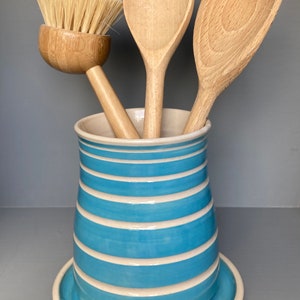 May include: A ceramic utensil holder with a light blue and white striped design, holding wooden spoons and a brush. The holder sits on a matching saucer. The spoons and brush are made of light-colored wood.