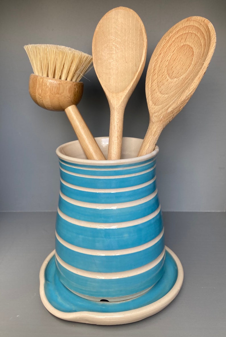 May include: A ceramic utensil holder with a light blue and white striped design, holding wooden spoons and a brush. The holder sits on a matching saucer. The spoons and brush are made of light-colored wood.