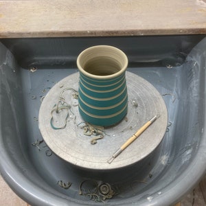 May include: A handmade ceramic vase in the process of being created on a pottery wheel. The vase is teal with horizontal stripes. A pottery tool rests on the wheel. The wheel is surrounded by a gray basin.