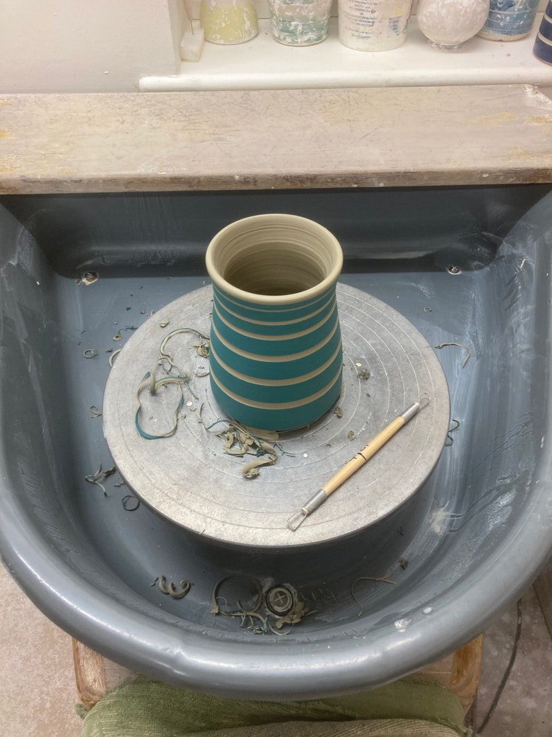 May include: A handmade ceramic vase in the process of being created on a pottery wheel. The vase is teal with horizontal stripes. A pottery tool rests on the wheel. The wheel is surrounded by a gray basin.