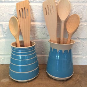 May include: Two blue and white ceramic utensil holders with wooden spoons and spatulas. One holder has horizontal stripes, the other has a dripping paint design. The kitchen tools are made of light-colored wood.