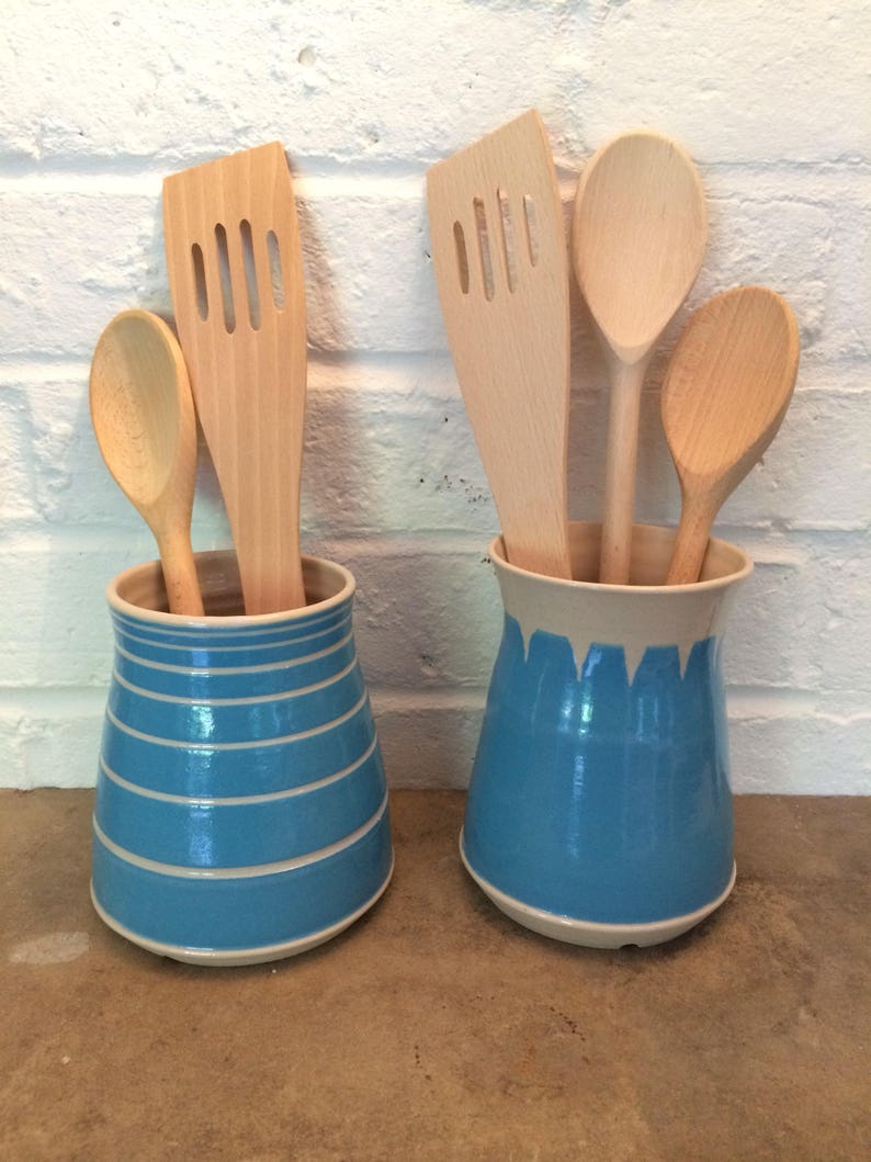 May include: Two blue and white ceramic utensil holders with wooden spoons and spatulas. One holder has horizontal stripes, the other has a dripping paint design. The kitchen tools are made of light-colored wood.