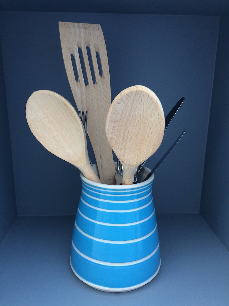 May include: A blue and white striped ceramic utensil holder filled with wooden kitchen tools. The tools include a slotted spatula, spoons, and a fork. The holder is cylindrical and sits on a blue surface.