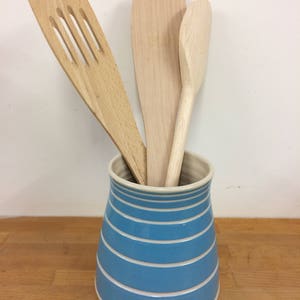 May include: A blue and white striped ceramic utensil holder with three wooden kitchen tools. The wooden tools include a slotted spatula, a flat spatula, and a spoon. The holder is cylindrical with horizontal stripes.