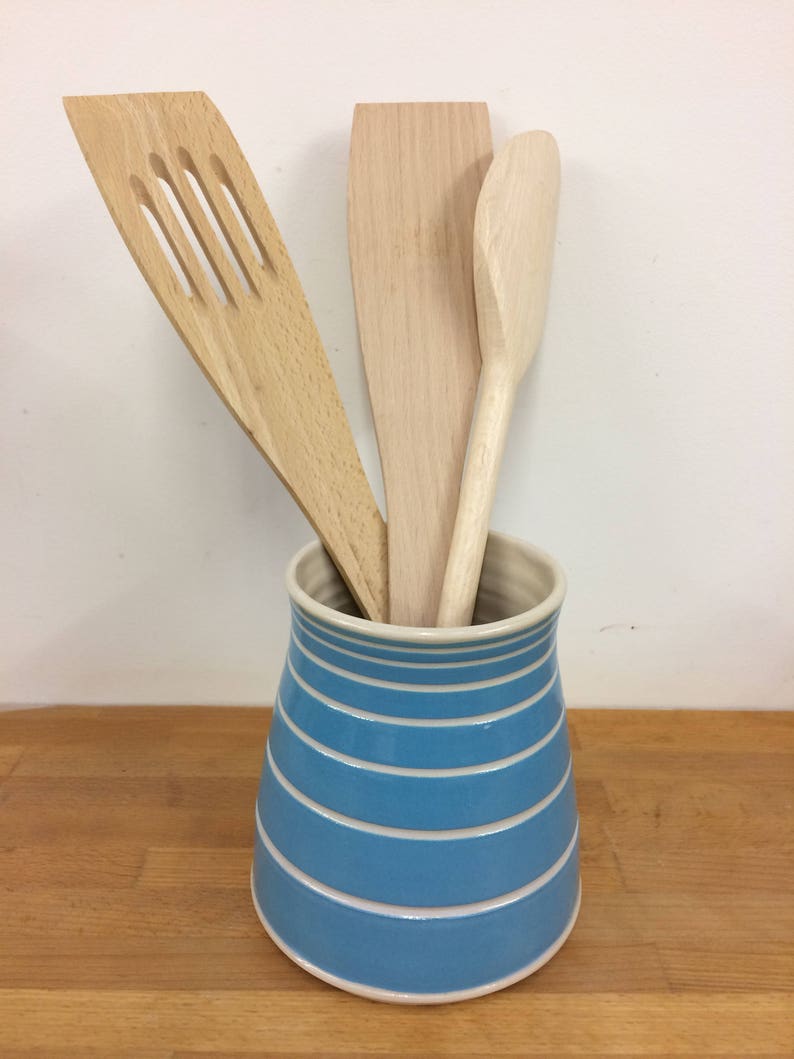 May include: A blue and white striped ceramic utensil holder with three wooden kitchen tools. The wooden tools include a slotted spatula, a flat spatula, and a spoon. The holder is cylindrical with horizontal stripes.