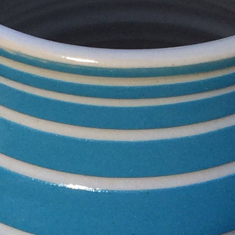 May include: Close-up of a ceramic mug with a white rim and alternating bands of white and turquoise blue. The bands are evenly spaced and create a striped pattern around the mug's exterior.