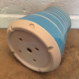 May include: A ceramic planter with a turquoise blue exterior and horizontal white stripes. The base is unglazed, with drainage holes and a stamped design. The planter has a slightly curved shape.