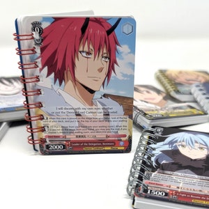 May include: Two small spiral bound notebooks with anime character illustrations on the covers. The notebook on the left features a red-haired character with horns and the text "I will discern with my own eyes whether or not the Demon Lord Carrion can be trusted." The notebook on the right features a blue-haired character with the text "Okay, Clayman, let's finish this."