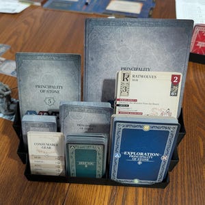 Puede incluir: Un organizador negro lleno de componentes de juegos de mesa. El organizador contiene cartas y tableros de juego con las etiquetas "Principality of Stone", "Ratwolves" y "Exploration Principality of Stone". Las cartas son de varios colores, incluyendo azul, verde y blanco.