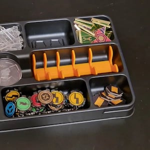 May include: A black plastic organizer tray with compartments for storing game pieces. The tray contains clear plastic tokens, small orange plastic dividers, and various colored game pieces with symbols.