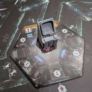 May include: A grey, 3D-printed miniature computer sits on a hexagonal game tile. The tile has a dark, detailed design with numbers and symbols. The computer has a screen and keyboard. The game piece is for a board game.
