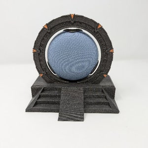 May include: A black 3D printed model of a Stargate, a fictional device from the TV show Stargate SG-1. The Stargate is a circular ring with a blue sphere in the center, and it is sitting on a black platform with steps.