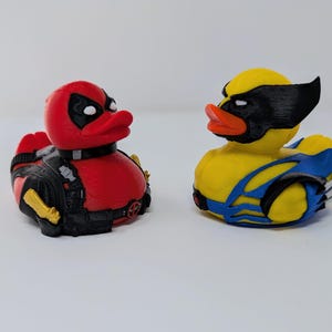 Puede incluir: Dos juguetes de pato de goma, uno rojo y negro con un diseño de Deadpool y el otro amarillo y azul con un diseño de Wolverine.