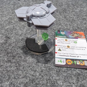 May include: A gray plastic spaceship miniature with a green base and a card that reads "CARRIERS" with the text "You may into Carrier Fleets for a cost of 1 per Fleet Power."  The card also has a +4 symbol with orange and gray icons.