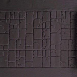 May include: A rolled-up, gray, textured mat with a cobblestone pattern. The mat is unrolled to reveal a section of the cobblestone design. The cobblestones are square and rectangular, with visible cracks between them. The overall effect is a realistic stone texture.