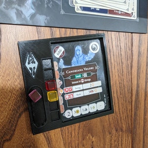 May include: A black plastic game character holder with a card showing a character named "Canoriana Velori" with a blue and white design. The card has the text "Sneak 3" and "Immune to damage." The holder has a red, yellow, and clear plastic dice and a purple plastic token.
