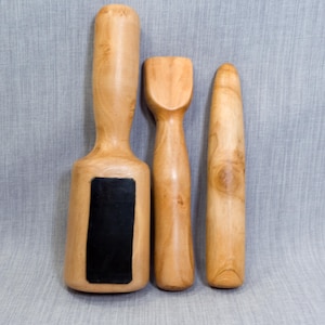 May include: Three light brown wooden tools with smooth, polished surfaces. The left tool is a mallet with a rectangular head and a black rectangular insert. The center tool has a flat, wide top, and the right tool is a tapered, cylindrical rod.