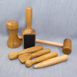 May include: A collection of six light brown wooden tools including a mallet with a black textured face, a rounded pestle-like tool, and four various shaped wooden burnishers or modeling sticks with smooth, polished surfaces and natural wood grain.