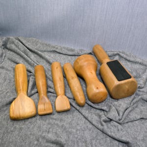 Könnte beinhalten: Eine Sammlung von sechs hellbraunen Holzwerkzeugen mit glatten, polierten Oberflächen. Das Set enthält verschiedene Formen wie flache Spatel, einen zylindrischen Stab, ein abgerundetes stößelartiges Werkzeug und einen rechteckigen Hammer mit schwarzer Gummiauflage.