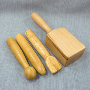 Op de afbeelding: Een verzameling van vier houten massagegereedschappen. De gereedschappen omvatten drie cilindrische vormen met handvatten en een rechthoekige hamer met een handvat. Het hout heeft een lichte, natuurlijke afwerking.
