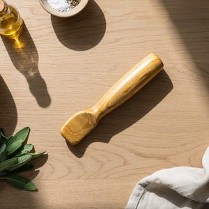 Peut inclure: Un pilon en bois avec une tête et une poignée arrondies repose sur une surface en bois clair. Un mortier et un pilon, une bouteille d'huile, un petit bol de sel et un bouquet de sauge sont également présents, suggérant un thème culinaire.