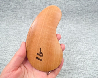 May include: A light brown wooden gua sha tool with a curved shape and a small engraved design. The tool is held in a hand.