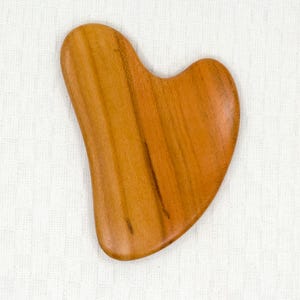 May include: A heart-shaped wooden gua sha tool, made of light brown wood with a smooth, polished surface. The tool has a slightly curved shape and is designed for use in facial massage.