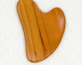 May include: A heart-shaped wooden gua sha tool, made of light brown wood with a smooth, polished surface. The tool has a slightly curved shape and is designed for use in facial massage.