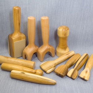 May include: A collection of ten light brown wooden massage tools with smooth, polished surfaces. The set includes various shapes such as cylindrical rollers, curved handheld massagers, a flat-bottomed block with a textured pad, and small paddles.