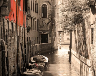 May include: A narrow canal in Venice, Italy, with several small boats docked along the sides. The canal is lined with old brick buildings, and the water is calm and reflective.