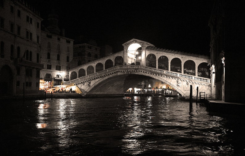 May include: A black and white photo of a bridge over a canal in Venice, Italy at night. The bridge is lit up and there are people walking across it. The water is reflecting the lights.