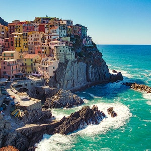 May include: A colourful village built on a cliff overlooking a turquoise ocean. The village is made up of many buildings with red, yellow, and orange roofs. The ocean is calm and the sky is blue.