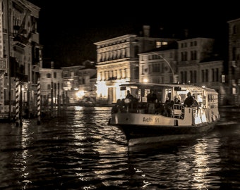May include: A black and white photo of a boat on a canal in Venice, Italy. The boat is a water taxi with the words "Actv 11" on the side. The boat is moving through the water and there are buildings on either side of the canal.