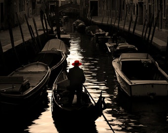 May include: A person in a red hat rows a gondola down a narrow canal in Venice, Italy.  The canal is lined with buildings and other boats.