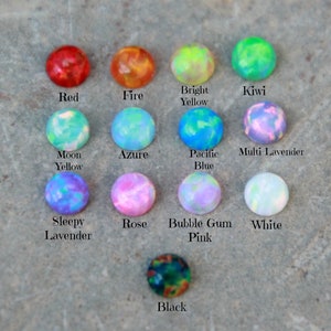 Op de afbeelding: Een verzameling ronde, kleurrijke opaal cabochons in verschillende kleuren, waaronder rood, oranje, groen, blauw en paars. Elke steen is gelabeld met zijn kleurnaam, zoals "Rood", "Vuur" en "Kiwi". De stenen zijn gerangschikt op een getextureerd, grijs oppervlak.