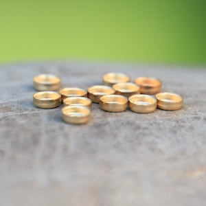 Puede incluir: Una colección de pequeñas cuentas circulares de color dorado. Las cuentas están espaciadas uniformemente y tienen una parte superior ligeramente cóncava. El fondo es una superficie gris neutra borrosa con un fondo verde.