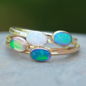 May include: Three gold rings with oval-shaped opal gemstones. The rings are stacked on top of each other. The opals are a variety of colours, including white, green, and blue.