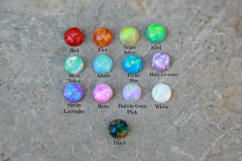 Op de afbeelding: Een verzameling ronde, kleurrijke opaal cabochons in verschillende kleuren, waaronder rood, oranje, groen, blauw en paars. Elke steen is gelabeld met zijn kleurnaam, zoals "Rood", "Vuur" en "Kiwi". De stenen zijn gerangschikt op een getextureerd, grijs oppervlak.