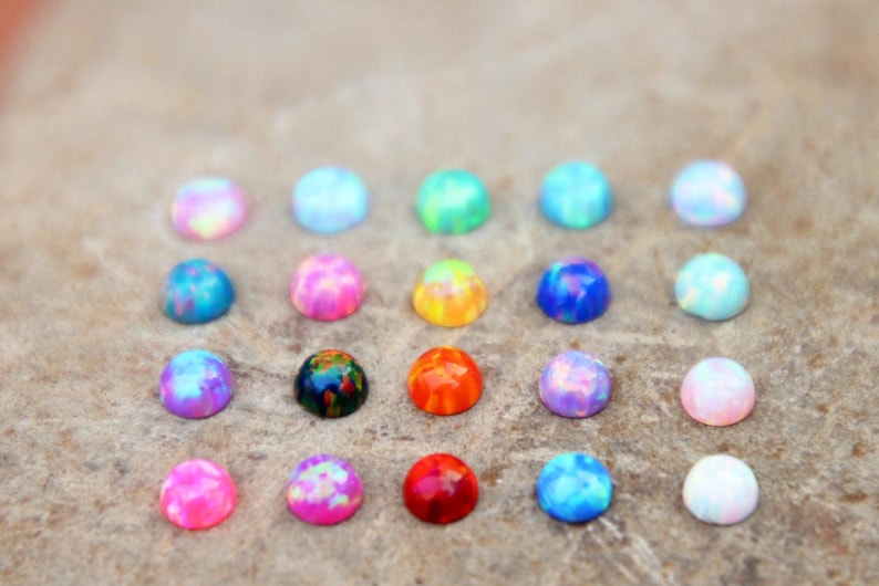 Op de afbeelding: Een verzameling kleurrijke, iriserende cabochons in verschillende tinten roze, blauw, groen en oranje. Deze kleine, gewelfde edelstenen zijn gerangschikt op een neutrale ondergrond, waarbij hun levendige, gespikkelde uiterlijk wordt getoond.