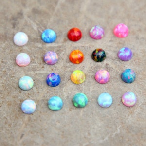 Op de afbeelding: Een verzameling kleurrijke, ronde opaal cabochons in verschillende tinten blauw, roze, rood, oranje, groen en paars. De opalen zijn gerangschikt op een neutrale, getextureerde ondergrond, die hun iriserende kwaliteiten benadrukt. Deze edelstenen zijn ideaal voor het maken van sieraden.