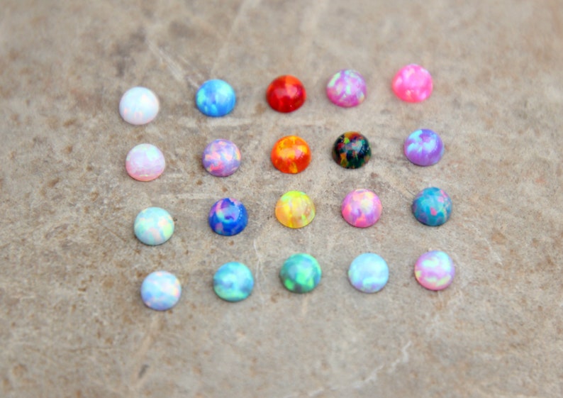 Op de afbeelding: Een verzameling kleurrijke, ronde opaal cabochons in verschillende tinten blauw, roze, rood, oranje, groen en paars. De opalen zijn gerangschikt op een neutrale, getextureerde ondergrond, die hun iriserende kwaliteiten benadrukt. Deze edelstenen zijn ideaal voor het maken van sieraden.