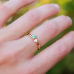 Op de afbeelding: Een ros&eacute;gouden ring met twee kleine edelstenen, een groene en een witte. De ring zit om een vinger, met een vage groene en gele achtergrond.
